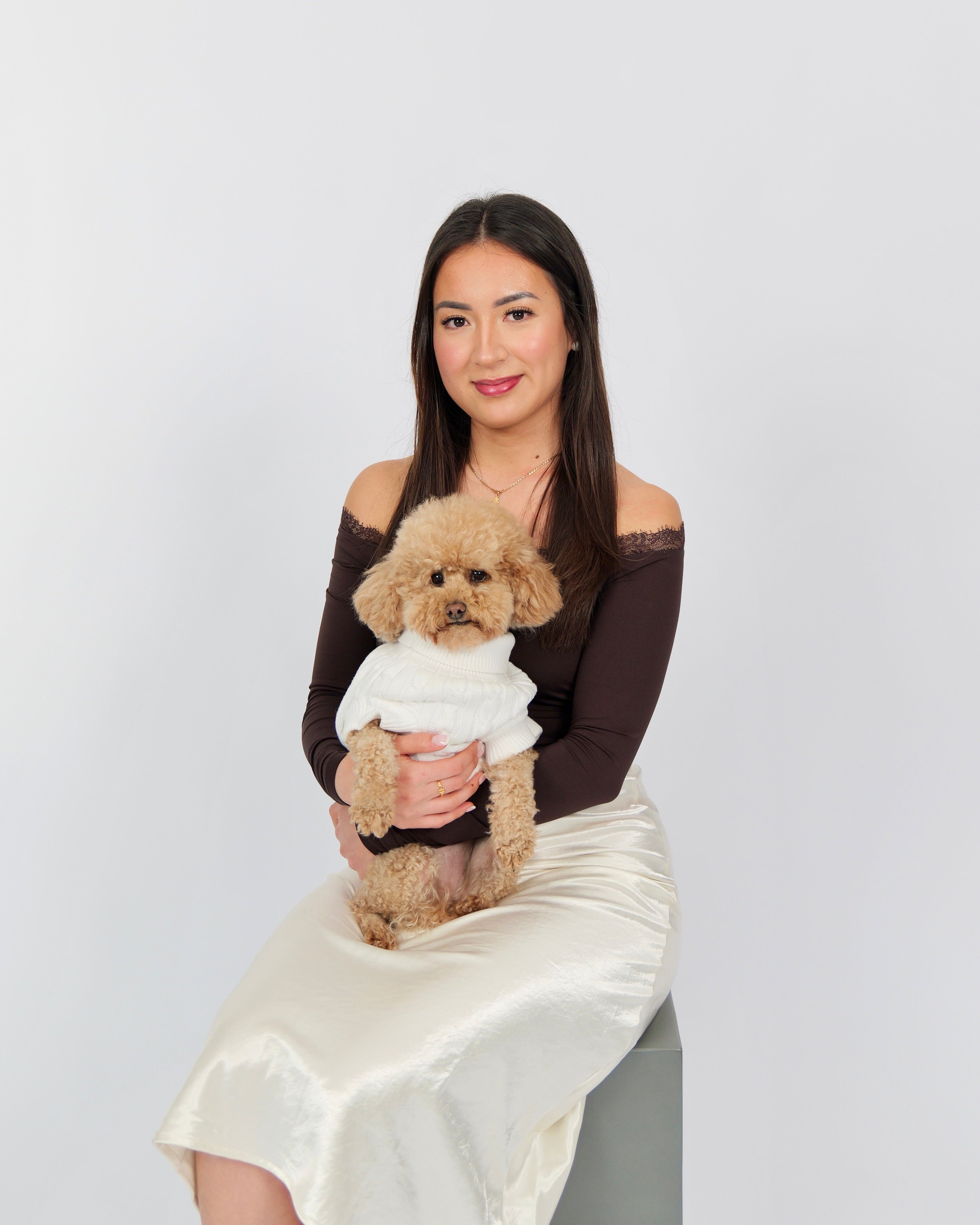 A woman with long dark hair sits on a gray stool against a light gray studio background, holding a small fluffy tan dog. The woman wears an off-the-shoulder dark brown top and a cream satin skirt, while the dog is dressed in a white sweater and black pants.